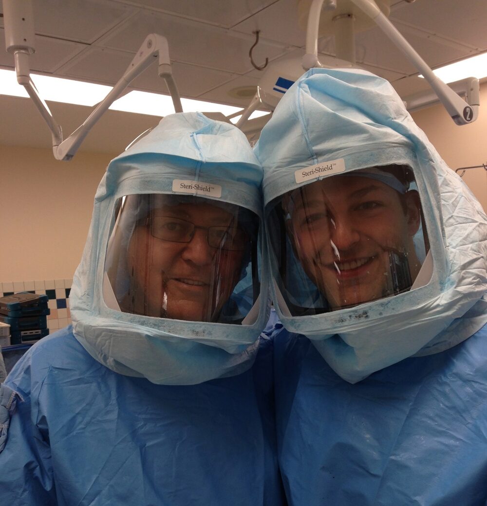 Dr. Matthew Wichman with his father, both gowned in surgical hoods in the operating room